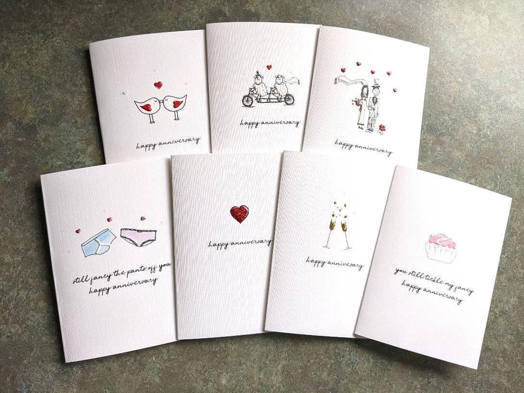 Seven white anniversary cards with hand-drawn illustrations arranged on a grey surface.