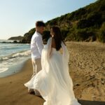 Couple in white wedding attire standing on a sandy beach by the ocean.