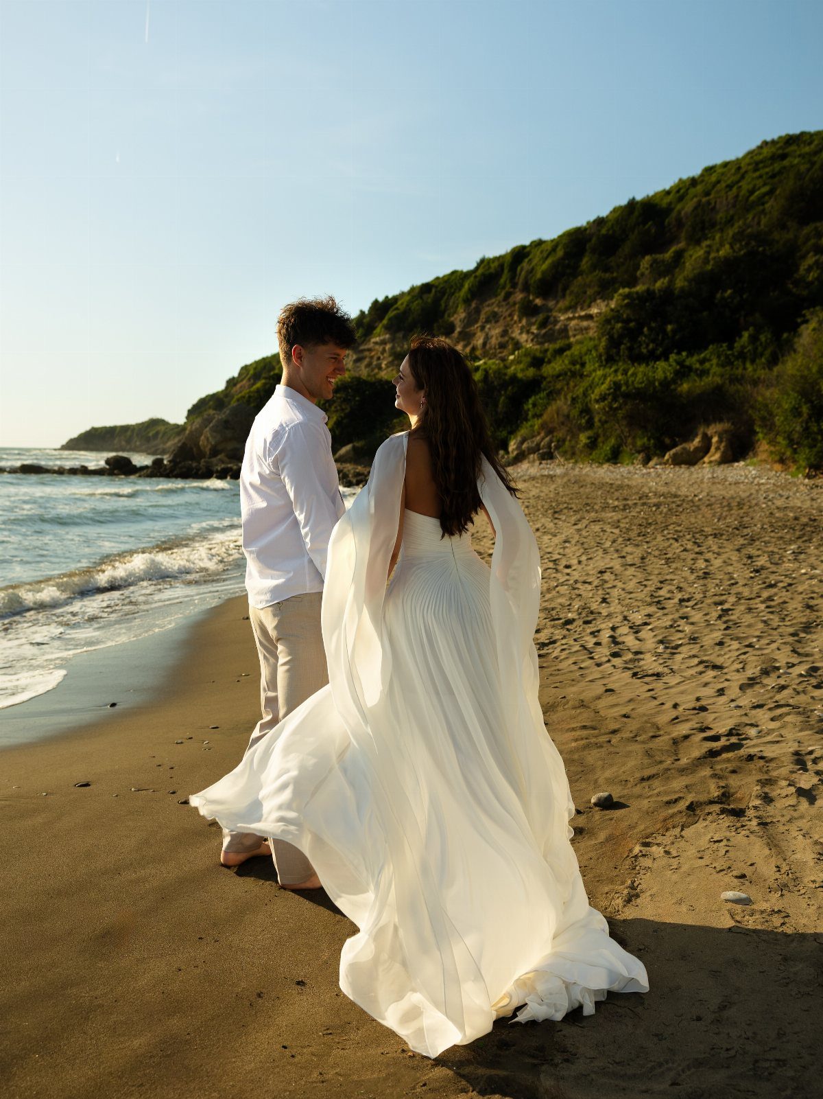 Couple in white wedding attire standing on a sandy beach by the ocean.