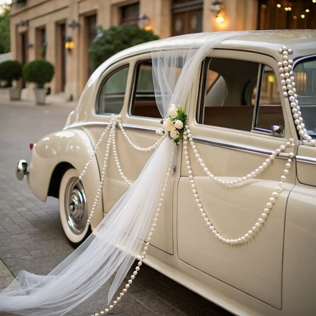 Cream vintage car decorated with white pearls, tulle, and flowers for a wedding.