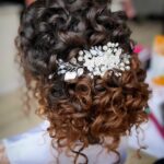 Curly bridal updo with dark brown and reddish hair featuring a pearl hairpiece.