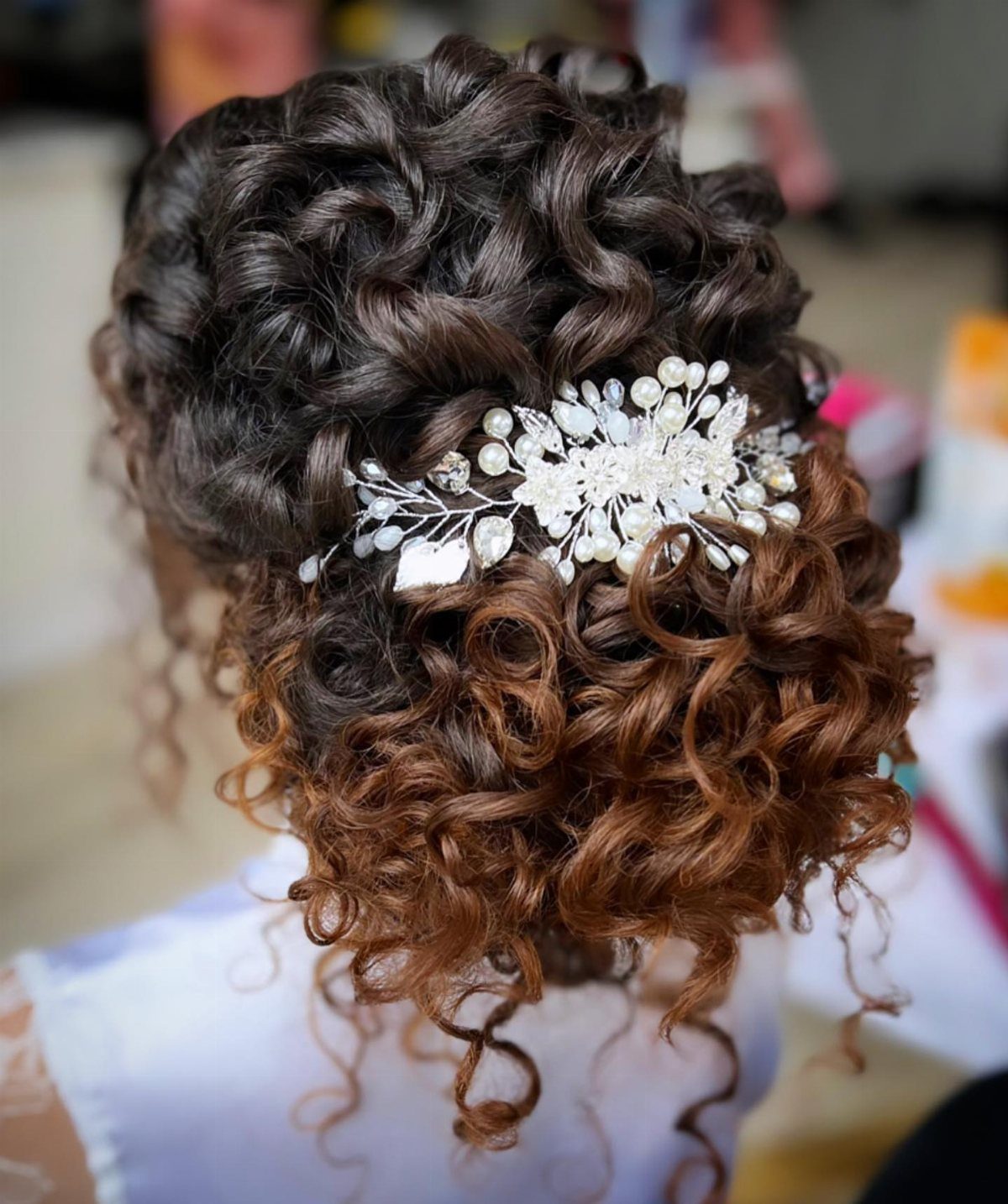 Curly bridal updo with dark brown and reddish hair featuring a pearl hairpiece.