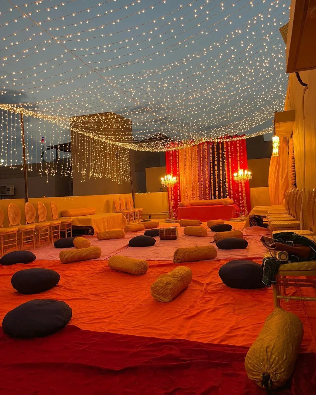 Outdoor mehndi decoration with orange flooring, cushions, and a canopy of fairy lights.