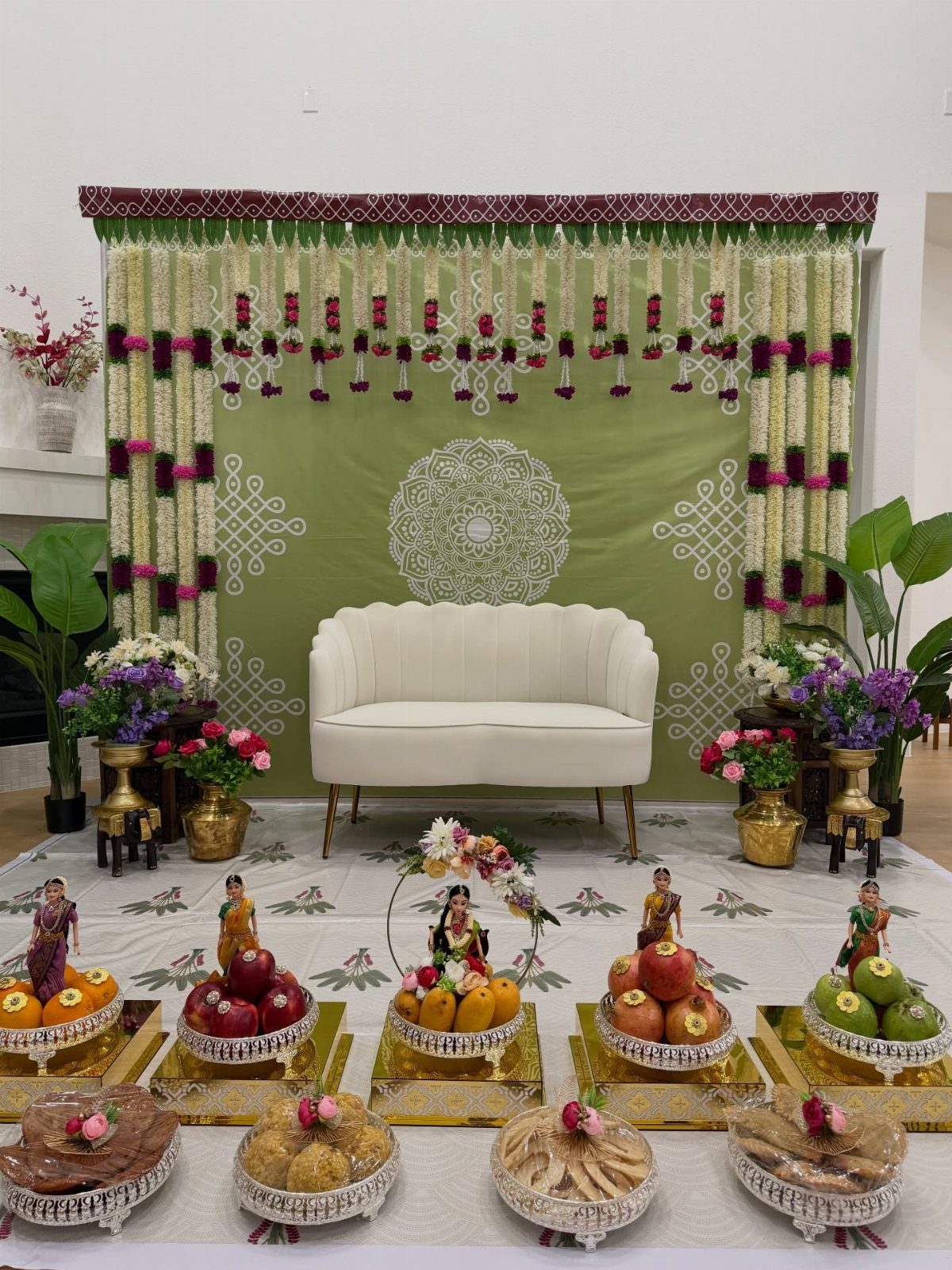 Festive decoration with a white sofa, green backdrop, floral garlands, and fruit platters.