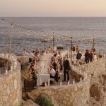 Long dinner table on a stone terrace by the ocean with guests and string lights.