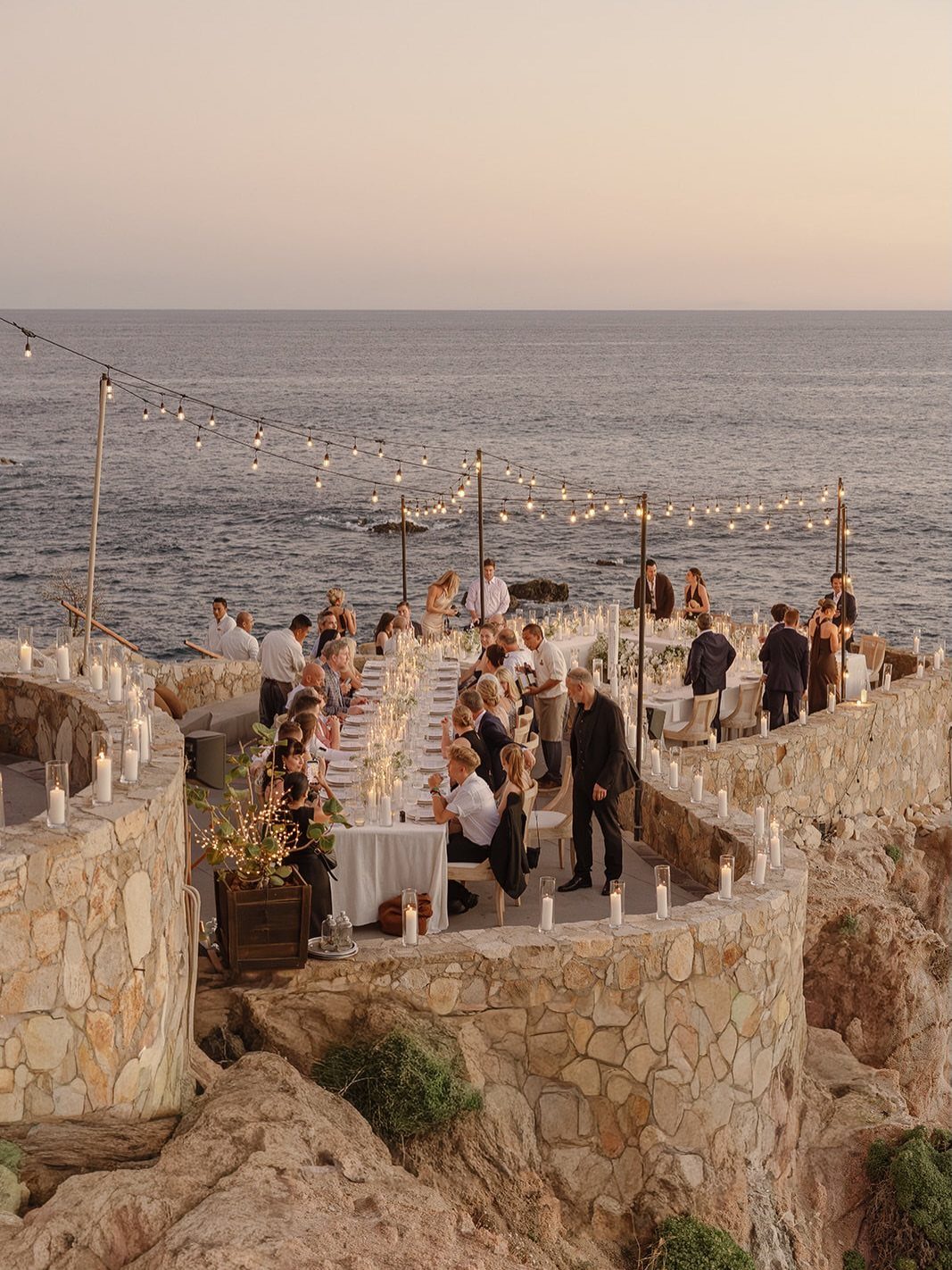 Long dinner table on a stone terrace by the ocean with guests and string lights.