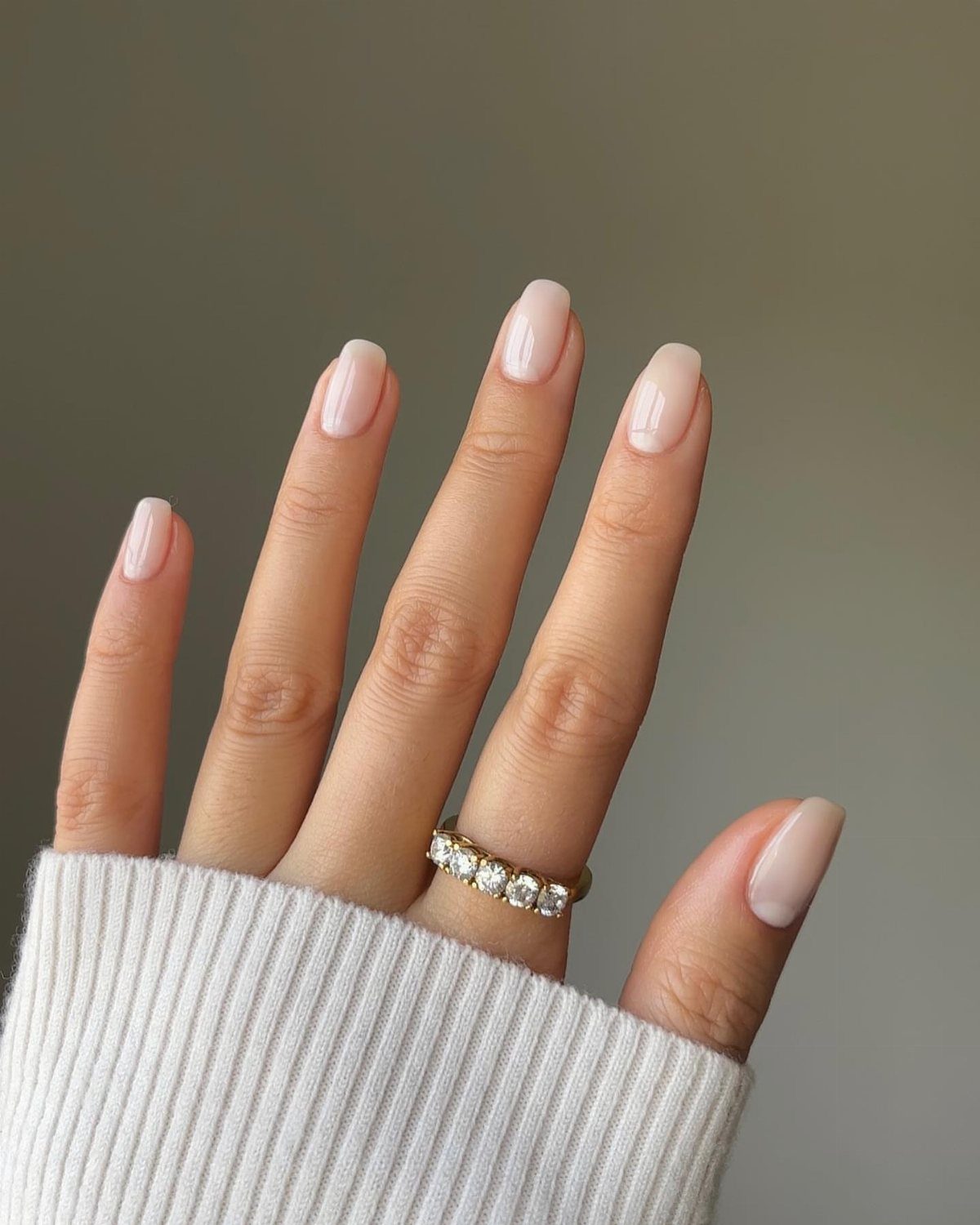 Hand with natural nude-pink almond nails and a diamond ring wearing a white sweater.