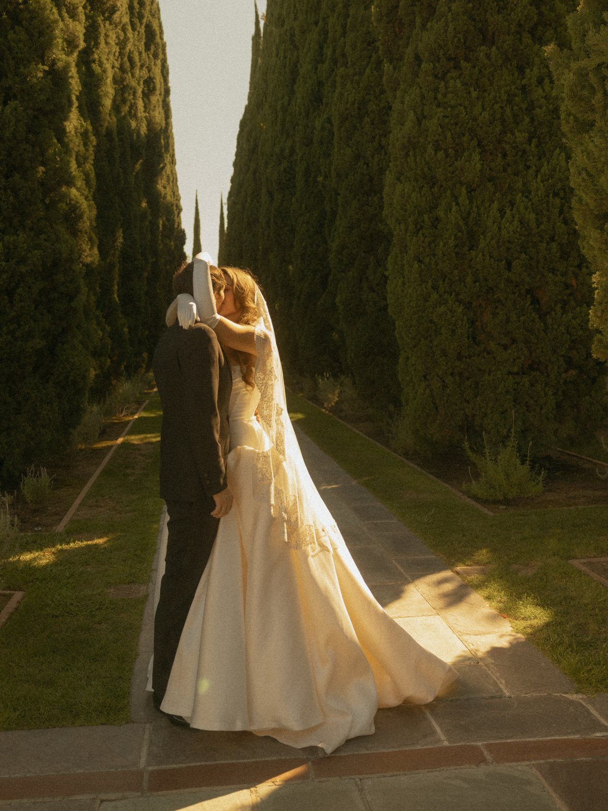 Bride and groom embracing and kissing on a path lined with cypress trees.